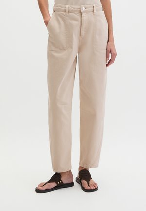 Pantalon beige taille haute à jambes droites avec poches plaquées à l'avant, porté avec des sandales marron foncé à enfiler, vu de la taille aux pieds.