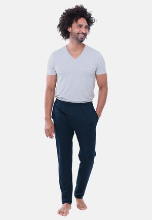 Mann mit lockigem Haar, der lächelt, trägt ein hellgraues kurzärmliges V-Ausschnitt-Shirt und dunkelblaue Hose, geht barfuß mit den Händen in den Taschen.