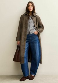 Brun och kräm houndstooth-kappa över grå knäpptröja, i kombination med blå vida jeans och brun handväska, mot en neutral bakgrund.