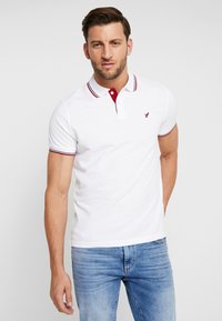 Polo bianco in cotone, con colletto rosso a strisce bianche e un piccolo logo ricamato sul petto. Maniche corte.