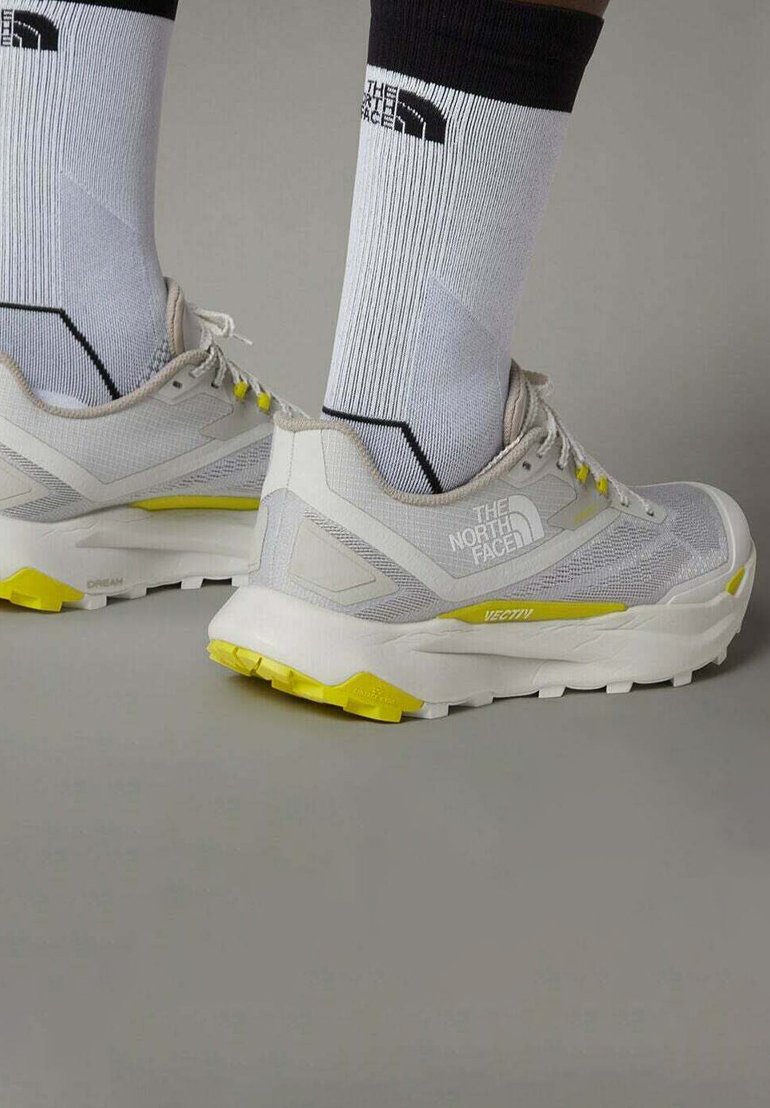 Lichtgrijze sportschoenen met gele accenten, ademend mesh bovenwerk en gestructureerde zool, gecombineerd met wit en zwart gestreepte sokken.