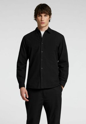 Camisa negra de manga larga con cierre de botones en el frente, corte ajustado y textura suave. Combinada con pantalones negros para un look coordinado.