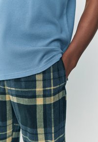 Hellblaues, geripptes Kurzarm-Oberteil, kombiniert mit dunkel karierten, strukturierten Hosen, die gelbe und grüne Linien aufweisen. Eine Tasche ist an den Hosen sichtbar.
