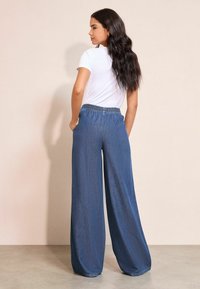 Bőszárú farmernadrág elasztikus derékpánttal; közép kék árnyalattal és puha textúrával; egy testre simuló fehér pólóval párosítva.