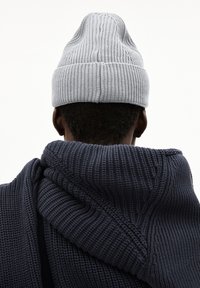 Persoon gezien vanaf de achterkant met een lichtgrijze gebreide beanie en een donkerblauwe chunky gebreide trui met een capuchon.