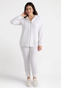 Felpa bianca con zip e cappuccio regolabile, due tasche frontali, abbinata a pantaloni slim-fit coordinati con spaccature laterali, indossata con sneaker bianche.