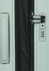 Hellgrüner Hartschalenkoffer mit schwarzem Reißverschluss, doppelten Metallverschlüssen und einem Kombinationsschloss. Verfügt über einen seitlichen Griff für einfaches Tragen.