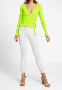 Neongrön långärmad omslagstopp med djup V-ringning, kombinerad med vita skinny jeans och nude band sandaler. Slät tyg, figurnära design.