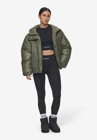 Olijfgroene pufferjack met een hoge kraag, knoopdetails en een glanzende textuur, gedragen over een zwarte crop top en leggins, gecombineerd met zwarte laarzen.