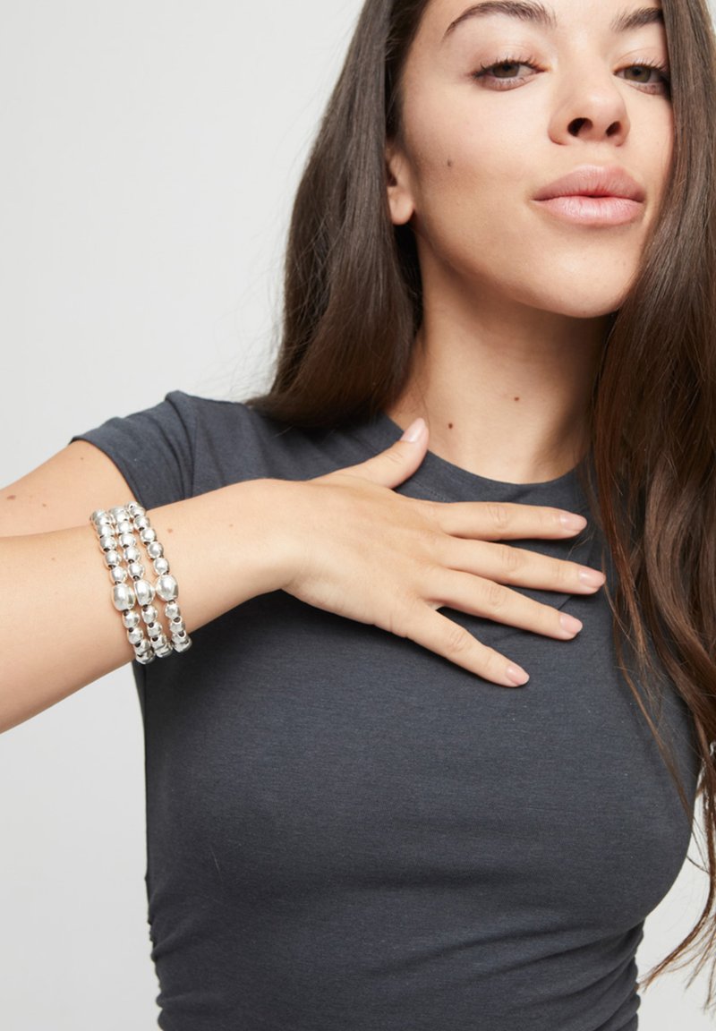 Pulsera de cuentas plateadas con tres vueltas, que presenta cuentas redondas y ovaladas, llevada en una muñeca con una camisa gris ajustada.