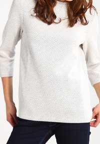 Suéter de punto texturizado en gris claro con un corte holgado, cuello redondo y mangas de tres cuartos, combinado con jeans oscuros.