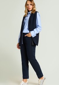 Donna in piedi che indossa una camicia azzurro chiaro con bottoni, un gilet smanicato scuro, jeans scuri e sneakers argentate su uno sfondo semplice.