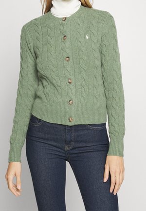Femme portant un cardigan vert en maille torsadée boutonné par-dessus un col roulé blanc, assorti à un jean bleu foncé.