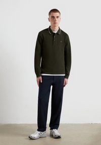 Langärmliges, dunkelgrünes Poloshirt mit weißen und schwarzen Kragenstreifen, kombiniert mit dunkelblauen Hosen und silbernen Sportschuhen. Einfaches Logo auf der Brust.