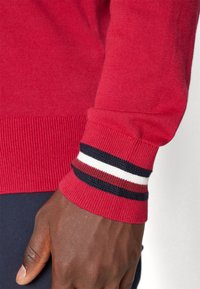 Pull rouge en tricot avec poignets côtelés présentant des rayures horizontales blanches et bleu marine. La texture est lisse et le design met en valeur une coupe ajustée.