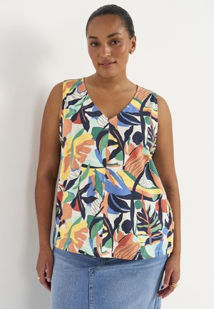 Kvinde iført en ærmeløs V-hals top med flerfarvet abstrakt bladmønster og lyseblåt denimskørt, stående mod hvid baggrund.