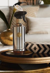 Sølvmetal sprayflaske med sort trigger, guld accenter, mærket 'White Basil Home Perfume', placeret på en guldbakke med dekorative genstande.