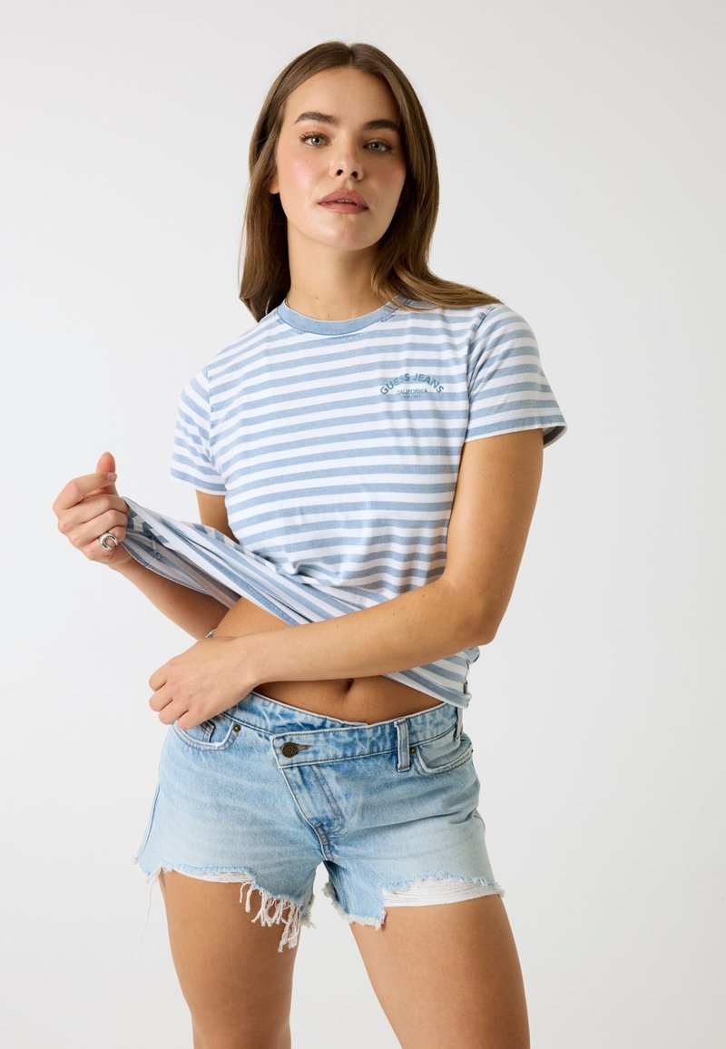 Jeune femme portant un t-shirt rayé bleu clair et un short en jean effiloché, soulevant légèrement son t-shirt pour révéler sa taille.