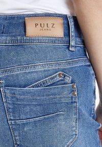 Jeans de mezclilla azul con una etiqueta de cuero en la cinturilla. Presenta dos bolsillos traseros, uno parcialmente visible, y detalles metálicos.