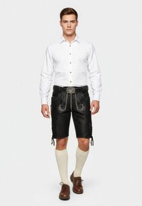 Camisa blanca de botones con pliegues, combinada con pantalones cortos de cuero negros con costuras amarillas y cordones laterales, completada con calcetines de punto color crema.