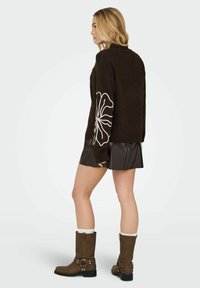 Pull en maille marron avec un motif floral sur la manche, associé à une jupe en cuir noire et des bottes marron. Texture lisse, style décontracté.