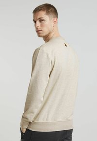 Felpa beige con scollo rotondo, maniche lunghe e un tessuto texturizzato. Dettaglio logo piccolo sul collo posteriore, abbinata a pantaloni scuri.