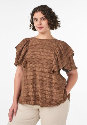 Kvinde med kort, krøllet hår iført en brun, tekstureret bluse med flæseærmer og lyse beige bukser, stående med den ene hånd i lommen.