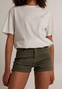 Camiseta de algodón blanca de manga corta con cuello redondo y un pequeño gráfico de sol. Combinada con shorts de mezclilla verdes con puños vueltas y bolsillos frontales.