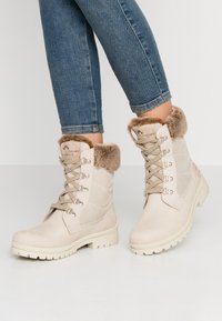 Beige ankelstøvler med falsk pelskant, suede materiale, metalløyer og snøring; robust såle og logodetalj på siden.