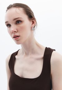 Top marrone senza maniche con scollatura quadrata; il modello indossa orecchini d'argento con un design ondulato su uno sfondo bianco semplice.
