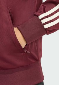 Maroon jacket featuring a zippered pocket and ribbed cuffs. White stripes accent the sleeve, highlighting its athletic design and texture.