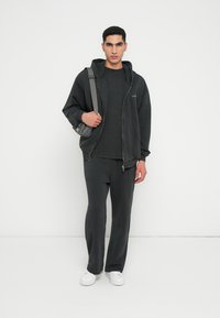 Sudadera negra con pantalones a juego, hechos de una tela suave. El modelo sostiene un bolso de bandolera gris. Zapatillas blancas completan el conjunto. Diseño simple y casual.