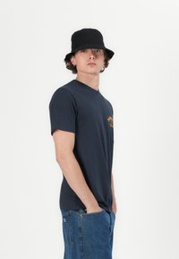 Navy short-sleeve t-shirt with a v-neck, featuring orange graphic text on the chest. Worn with blue denim jeans and a black bucket hat.