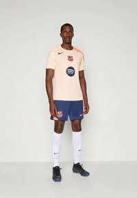 Gai bēša futbols kreklis ar oranžiem plecu akcentiem, Barselonas logo un Spotify druku. Tumši zilas šortus, baltas zeķes un melnus sporta apavus.