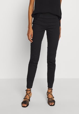 Woman wearing slim black pants and black strappy heeled sandals, standing against a plain light background.