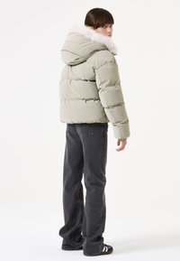 Khaki pufferjack met een nepbont capuchon, met een horizontaal gestikt ontwerp. G gedragen met donkergrijze jeans en zwarte sandalen.