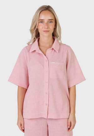Kvinne med blondt hår iført en rosa kortermet skjorte med knapper og matchende shorts foran en ensfarget lys bakgrunn.