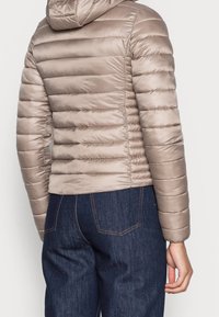 Beżowa ocieplana kurtka z kapturem, z poziomymi przeszyciami, o błyszczącej fakturze i dopasowanym kroju, w zestawie z ciemnoniebieskimi dżinsami.