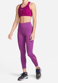 Fioletowy stanik sportowy i legginsy z wysokim stanem, oba wykonane z elastycznego materiału. Buty sportowe mają wielokolorowy wzór i czarne sznurówki.