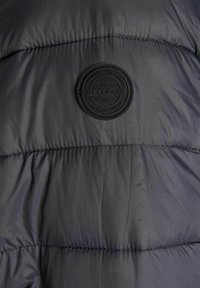 Puffer jacket in dark gray fabric with a quilted design. Features a textured, black circular logo patch and visible stitching details.