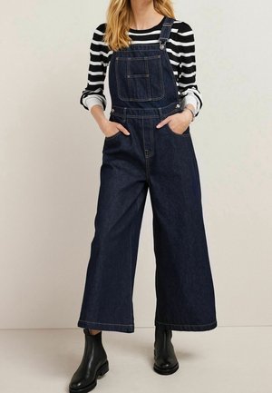 Nainen, joka pukeutunut tummansinisiin leveälahkeisiin farkkuhaalareihin, mustavalkoraidalliseen pitkähihnaiseen paitaan ja mustiin nilkkureihin.