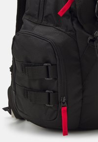 Black fabric backpack featuring a textured outer, multiple pockets with black straps, and a red zipper pull accent.