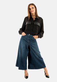 Blusa negra de botones con detalles de cuero, combinada con pantalones vaqueros de pierna ancha azules. La modelo lleva tacones altos negros y tiene las manos en los bolsillos.