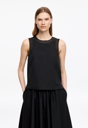 Femme aux cheveux raides portant un haut noir sans manches avec des panneaux transparents et une jupe noire, debout les mains dans les poches sur fond blanc.