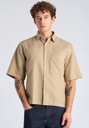 Junger Mann mit lockigem Haar, der ein beige kurzärmeliges Hemd mit Knöpfen und dunkelblaue Jeans trägt, und Tätowierungen auf beiden Unterarmen zeigt.