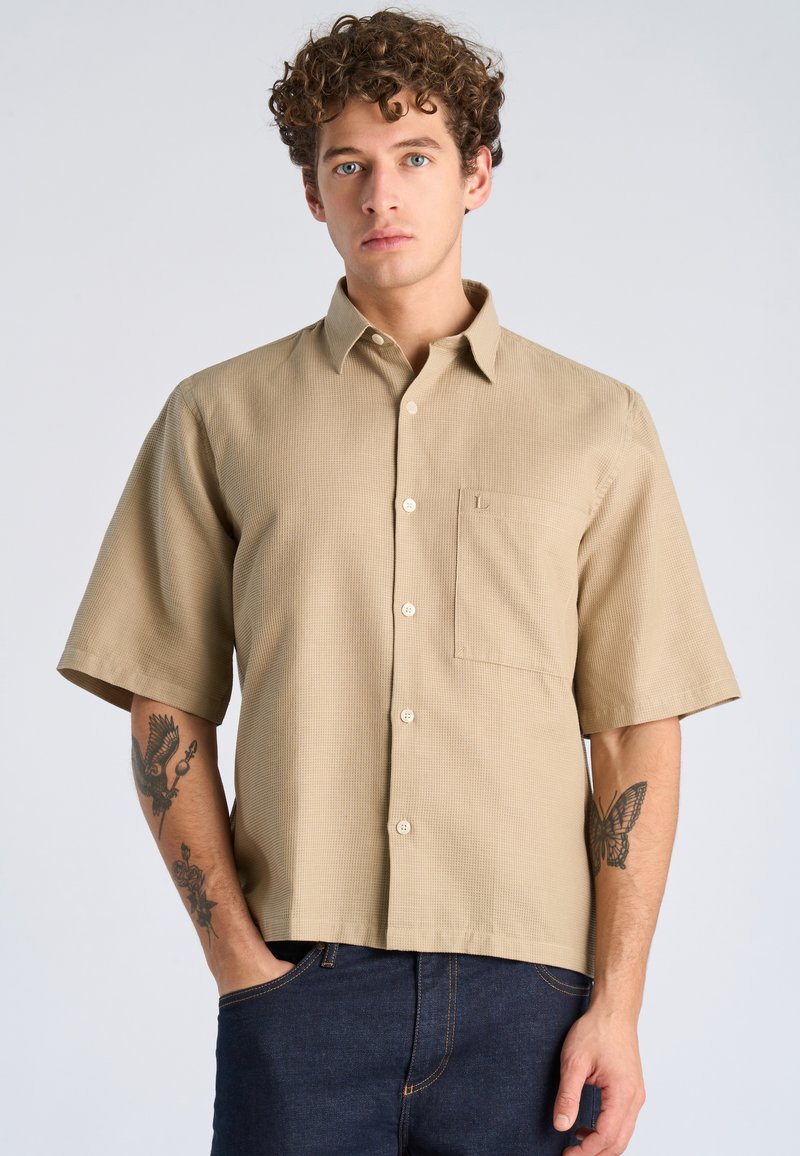 Jeune homme aux cheveux bouclés portant une chemise beige à manches courtes boutonnée et un jean bleu foncé, montrant des tatouages sur les deux avant-bras.