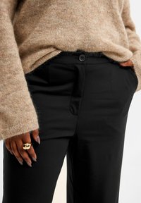 Zwarte broek met een gladde textuur, voorzien van een knoopsluiting en zakken, gecombineerd met een fluffy beige trui. Gouden ring om de hand.