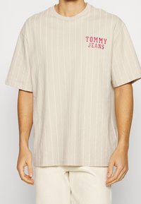 béžové tričko s krátkým rukávem s vertikálními bílými pruhy a červeným logem "TOMMY JEANS" na hrudi; vyrobeno z měkké bavlněné tkaniny.