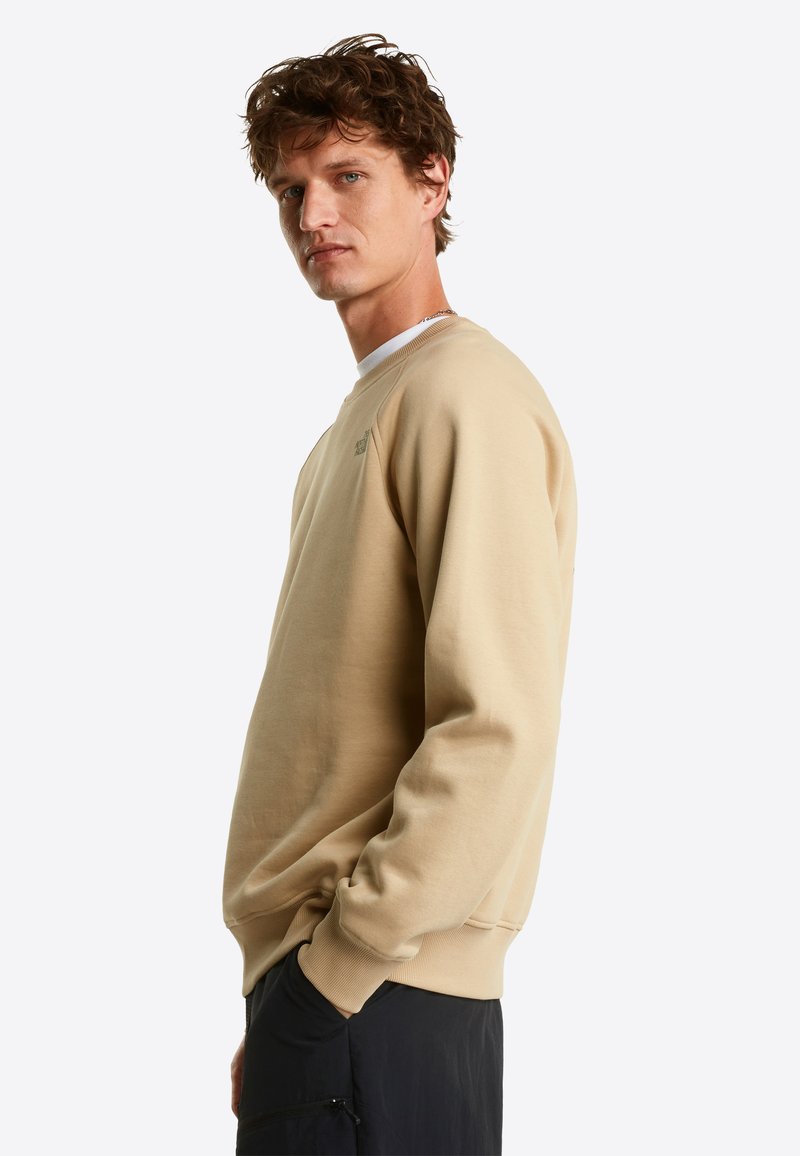 Man with curly hair wearing a beige sweatshirt and black pants, standing in profile with one hand in pocket against a white background.