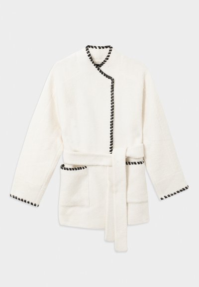 Peignoir blanc avec des finitions noires, doté d'une taille ceinturée, de manches longues et de deux poches latérales dans un tissu doux et texturé.